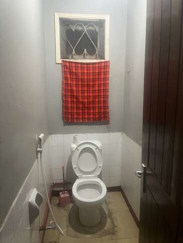 Il s'agit d'une petite salle de bains équipée d'un WC blanc, d'une brosse de toilettes, d'une douchette de bidet, de murs carrelés et d'une fenêtre recouverte d'un tissu à carreaux rouges.