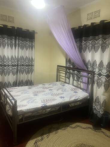 Un lit soigneusement fait, avec un drap à motifs et un cadre en métal, trône dans une chambre chaleureuse, décorée de rideaux à motifs, d'un tapis et d'une moustiquaire violette suspendue au-dessus.