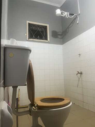 Cette image montre une salle de bains simple équipée de toilettes, d'un siège de WC en bois, de murs carrelés, d'une petite fenêtre, d'un robinet, d'une serpillière dans un coin et d'une pomme de douche apparente.