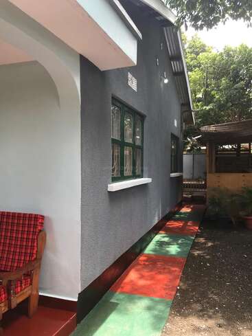 Cette image montre la façade d'une maison aux murs gris, aux encadrements de fenêtres verts et à l'allée pavée de carreaux rouges et verts. On y voit une chaise d'extérieur avec des coussins rouges.