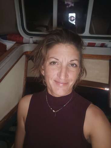 Una mujer con el pelo corto y revuelto y un piercing en la nariz sonríe con dulzura. Lleva una camiseta sin mangas y un collar, y posa en el interior de lo que parece ser un barco.