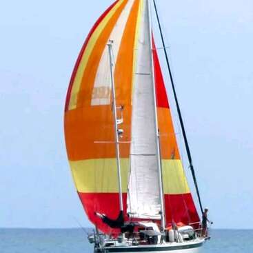 Un velero de vivos colores, con grandes velas naranjas, amarillas y rojas, se desliza sobre las tranquilas aguas azules bajo un cielo despejado, con varios pasajeros a bordo.