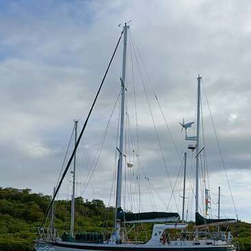 Ein Segelboot namens „Jubilate“ liegt vor Anker in der Nähe üppig grüner Hügel, begleitet von einem kleinen Boot und einem Kajak, unter einem teilweise bewölkten Himmel auf ruhigem Wasser.