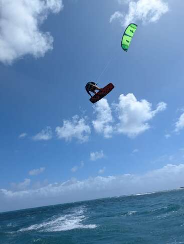 Un kitesurfista surca las alturas sobre el océano, impulsado por una cometa de color verde brillante bajo un cielo azul intenso, rodeado de nubes blancas y esponjosas y olas agitadas.