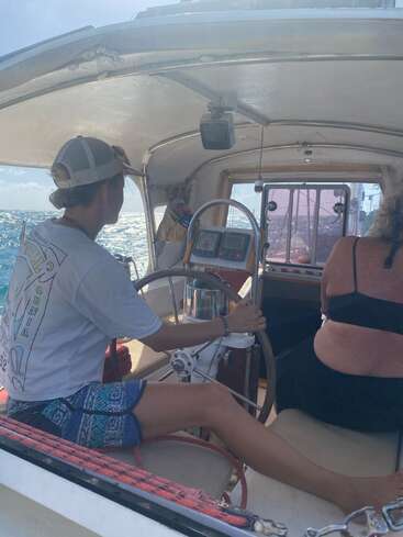 Hay dos personas en un barco. Una lleva el timón, concentrada en navegar por mar abierto, mientras que la otra está sentada, relajada, disfrutando del sol.
