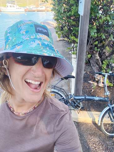Una persona sonriente lleva un sombrero de pescador de colores, gafas de sol y un collar de cuentas, y posa junto a una bicicleta a orillas del agua, con barcas y vegetación al fondo.