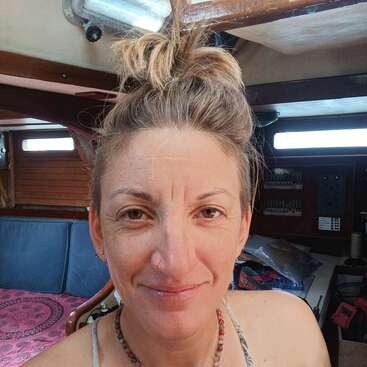 A woman with a nose ring and beaded necklace smiles gently inside a wooden boat cabin, with sunlight streaming through the windows and various items behind her.