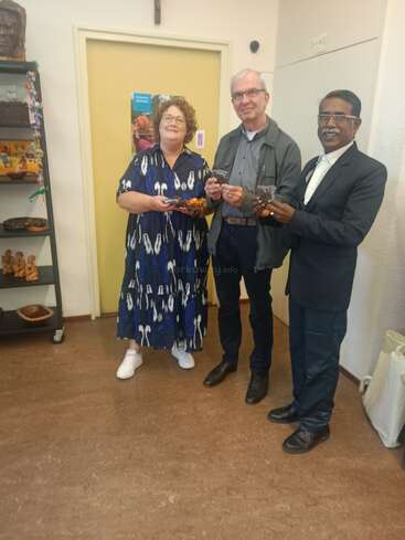 Three people stand together indoors, smiling and holding small objects. Behind them is a yellow door, shelves with decorative items, and artwork on the wall.