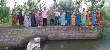 A group of women and two foreign visitors stand smiling on a stone platform above a water body, surrounded by lush greenery and colorful sarees.