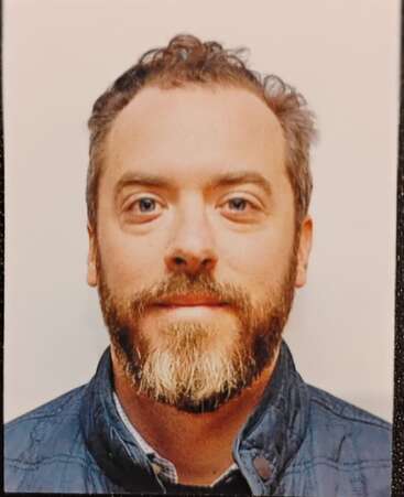 Un homme aux cheveux courts bouclés et à la barbe, vêtu d'une veste matelassée bleue, regarde directement l'appareil photo sur un fond uni de couleur claire, l'expression est neutre.