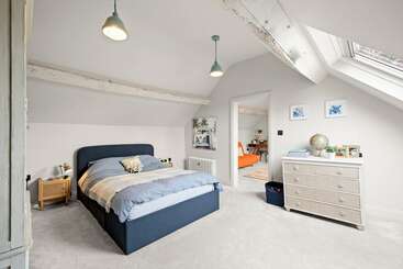 Une chambre lumineuse avec un lit bleu, des murs blancs, des poutres apparentes, une lucarne, des lampes suspendues, une commode, et une pièce adjacente confortable avec des meubles orange.