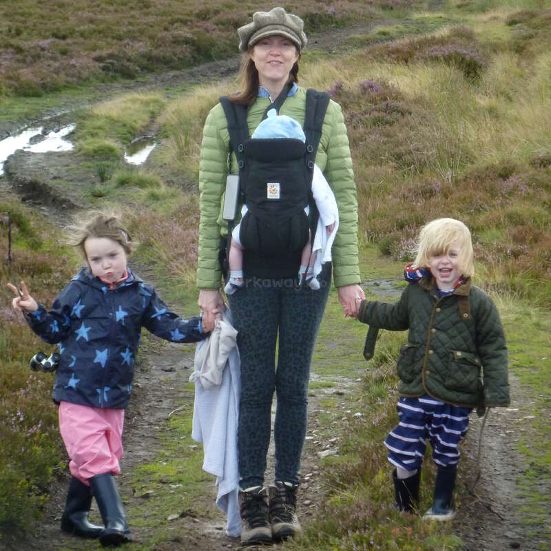 Une femme se tient en plein air sur un chemin boueux, portant un bébé dans un porte-bébé avant et tenant la main de deux enfants souriants portant des vêtements de plein air.
