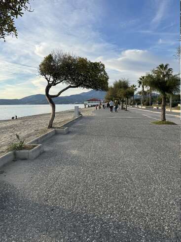 Un tranquilo paseo marítimo con árboles, gente paseando, una playa de arena a la izquierda, aguas tranquilas, montañas lejanas y un cielo azul brillante.