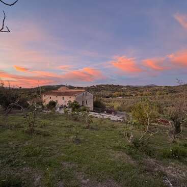 Una casa rústica se asienta apaciblemente sobre una colina cubierta de hierba, rodeada de árboles y un paisaje ondulado bajo un hermoso cielo pintado de nubes naranjas y rosas al atardecer.