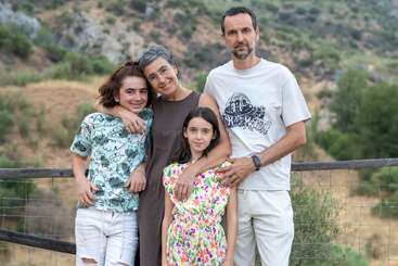 A family of four stands close together, smiling outdoors. They are surrounded by greenery and hills, posing near a wooden fence. The scene feels warm and loving.