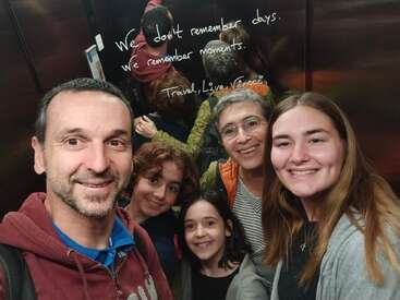 Un grupo de cinco personas sonríen juntas en un ascensor, posando para un selfie. Un espejo detrás de ellos muestra una cita inspiradora sobre cómo recordar momentos.