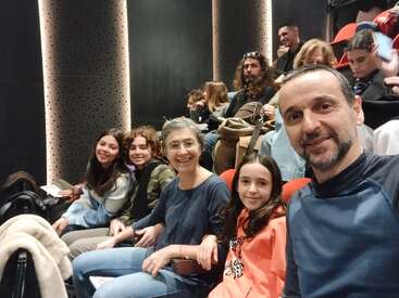 Una familia está sentada junta en un teatro, sonriendo a la cámara. El público detrás de ellos parece interesado, esperando a que comience una actuación o un evento.