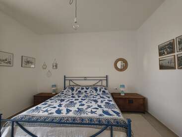 This is a minimalist bedroom with a blue metal bed frame, sea-themed bedspread, wooden nightstands, wall art, a round mirror, and simple decor.