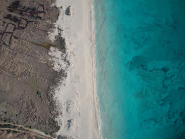This aerial view shows a white sandy beach bordered by turquoise ocean on the right and dry, rugged, brown terrain with ruins on the left. Peaceful contrast.