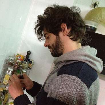Das Bild zeigt einen Mann mit dunklen Haaren und einem Bart, der einen grauen Pullover trägt und vor einem Gewürzregal steht, in dem er ein Messer und eine Flasche hält.