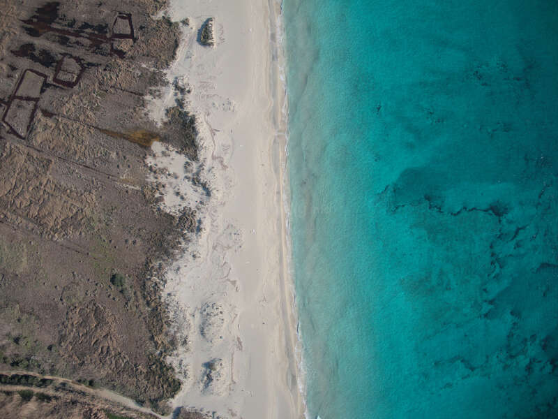 This aerial view shows a white sandy beach bordered by turquoise ocean on the right and dry, rugged, brown terrain with ruins on the left. Peaceful contrast.