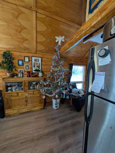 Im Inneren der gemütlichen Holzhütte gibt es einen geschmückten Weihnachtsbaum, einen Hund, der auf einer Couch liegt, Familienfotos an der Wand und einen Kühlschrank mit Notizen.