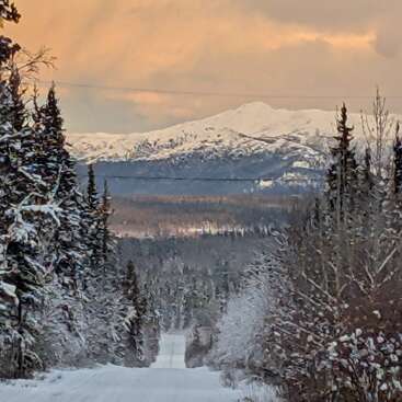 Uma estrada coberta de neve se estende por uma floresta de árvores geladas, levando a montanhas distantes e majestosas sob um céu dramático e tingido de laranja ao pôr do sol. Cena serena de inverno.