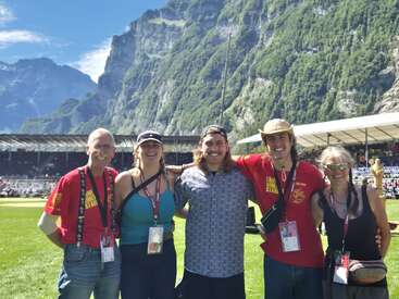Fünf lächelnde Menschen posieren zusammen in einem Stadion, im Hintergrund erheben sich die Berge und der sonnige Tag unterstreicht die festliche, lebhafte Atmosphäre mit den versammelten Menschenmengen.