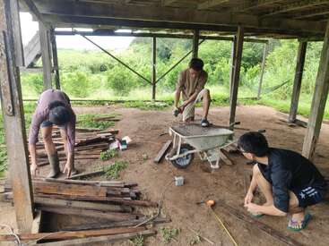 Três pessoas estão trabalhando sob uma estrutura de madeira, manuseando tábuas e ferramentas. Um carrinho de mão e uma fita métrica estão por perto, rodeados por vegetação e uma paisagem aberta.