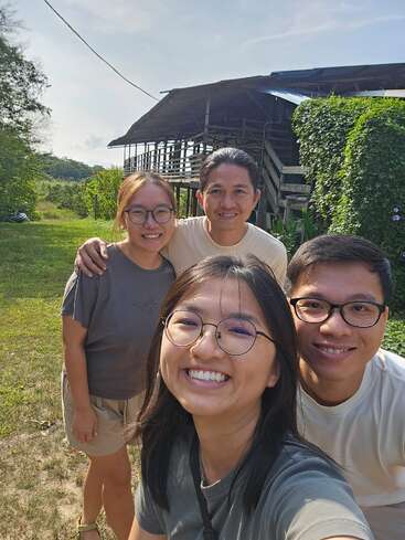 Quatro amigos sorridentes tiram uma selfie ao ar livre num dia ensolarado, em pé sobre a grama verde, com uma casa rústica de madeira e uma vegetação exuberante ao fundo.