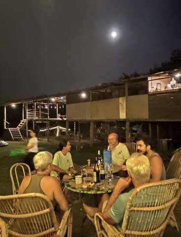 Sob uma lua brilhante, seis pessoas estão sentadas ao redor de uma mesa ao ar livre, curtindo bebidas e conversando perto de uma casa rústica e bem iluminada à noite. Um ambiente aconchegante.