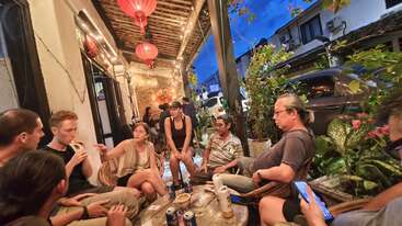 Um grupo de pessoas está sentado junto do lado de fora de um café aconchegante, sob lanternas penduradas, à noite, rodeado por plantas, bebidas e conversas animadas, curtindo a companhia um do outro.