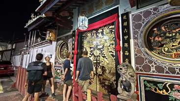 Quatro pessoas observam à noite a entrada de um templo ricamente decorada, com dragões dourados, entalhes intricados, lanternas, detalhes em vermelho e elementos arquitetônicos e símbolos tradicionais chineses.