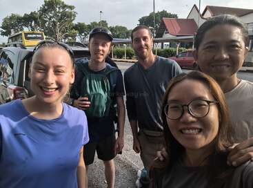 Cinco amigos sorridentes estão ao ar livre, posando juntos para uma selfie. Há carros, árvores e prédios ao fundo. O clima é alegre e descontraído.