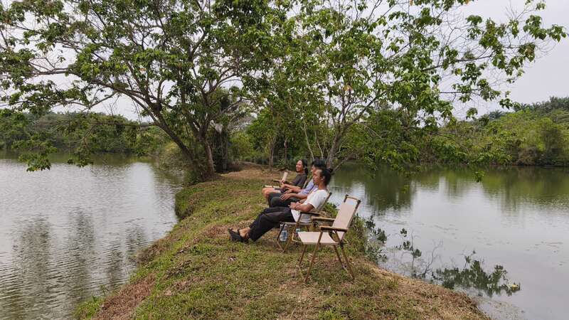 Três pessoas estão sentadas em cadeiras dobráveis à beira de um rio tranquilo, rodeadas por árvores, relaxando e curtindo a natureza. O clima é calmo e sereno, perfeito para descontrair.