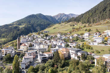 Ce charmant village de montagne se caractérise par des bâtiments blancs groupés, un clocher proéminent, une végétation luxuriante et des collines boisées, le tout entouré de hautes montagnes alpines pittoresques. Atmosphère paisible.