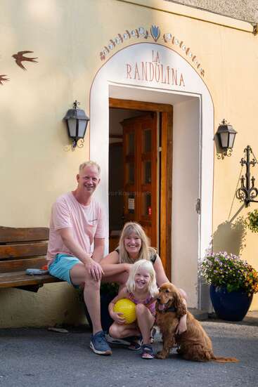 Une famille souriante de trois personnes, avec une jeune fille tenant un ballon jaune et un chien marron, pose joyeusement devant le bâtiment de "La Randulina". Des fleurs décorent l'entrée.