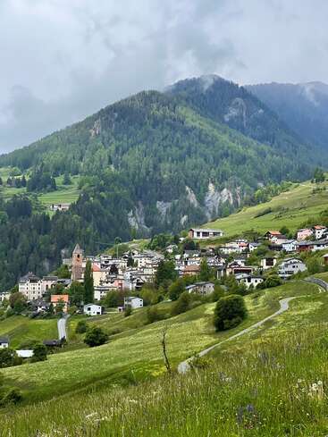 Un village pittoresque aux maisons colorées est niché sur une colline verdoyante, entouré de forêts denses et de montagnes imposantes, sous un ciel nuageux et brumeux. Paysage paisible et pittoresque.