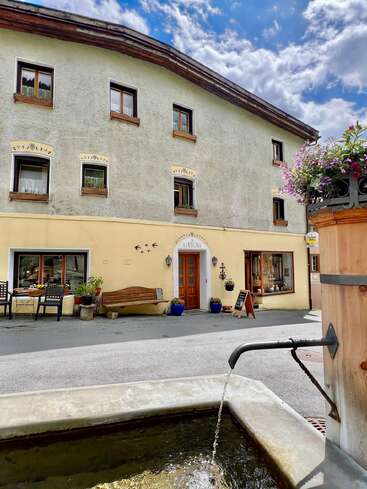 Un charmant bâtiment européen avec des fenêtres rustiques, une porte en bois, des sièges en plein air, des pots de fleurs et une fontaine en pierre par une journée lumineuse et partiellement nuageuse.