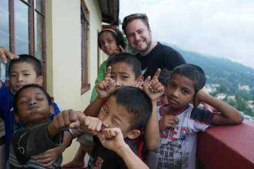 The image depicts a man posing with a group of children on a balcony, set against a scenic backdrop of a mountainous landscape.
