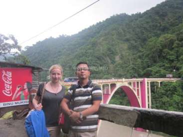 The image depicts a man and woman standing in front of a bridge, with a Coca-Cola advertisement and a mountainous background. The man is wearing a striped shirt.