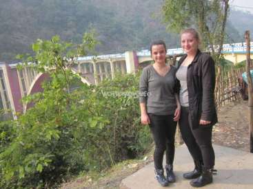 The image depicts two women standing on a path in front of a bridge, with a serene mountainous backdrop and lush greenery.