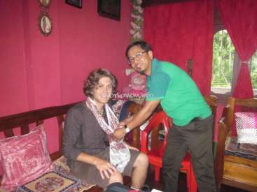 The image depicts two men in a room with pink walls, one seated on a wooden bench and the other standing beside him, shaking hands.
