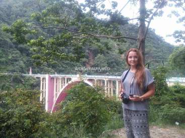The image depicts a woman standing in front of a bridge, surrounded by lush greenery and trees, with a serene and natural atmosphere.