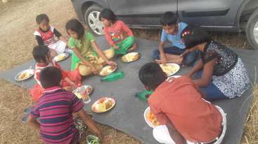 The image shows a group of children sitting on the ground, likely at a picnic or outdoor event, with a table and chairs in the background, surrounded by a natural setting.