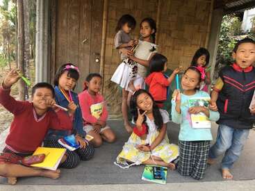A group of smiling children pose cheerfully with books and pens outside a simple bamboo house, expressing joy and togetherness in a rural setting.