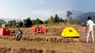 Four people are camping in a dry field surrounded by trees and mountains. Three colorful tents are pitched, and the sky is clear with light clouds.