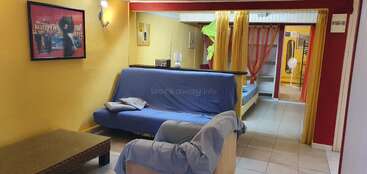 A cozy living space with yellow and red walls, blue futons, a coffee table, wall art, bright curtains, beds, and tiled floor, creating a vibrant atmosphere.