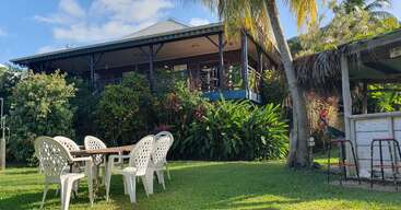 A cozy tropical house with a veranda is surrounded by lush greenery and palm trees. White plastic chairs and a table sit on a grassy lawn.
