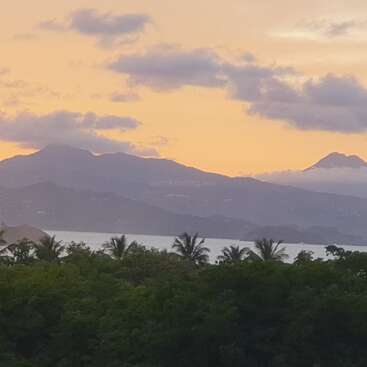 A serene sunrise or sunset scene with a golden sky, misty mountains, palm trees, calm water, and lush greenery in the foreground beneath scattered clouds.